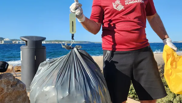 Voluntario pesando una bolsa de basura grande con una báscula de mano junto al mar