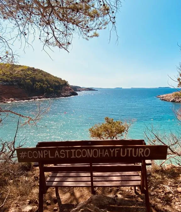 Banco de madera con el cartel #ConPlasticoNoHayFuturo frente a una cala de aguas turquesas